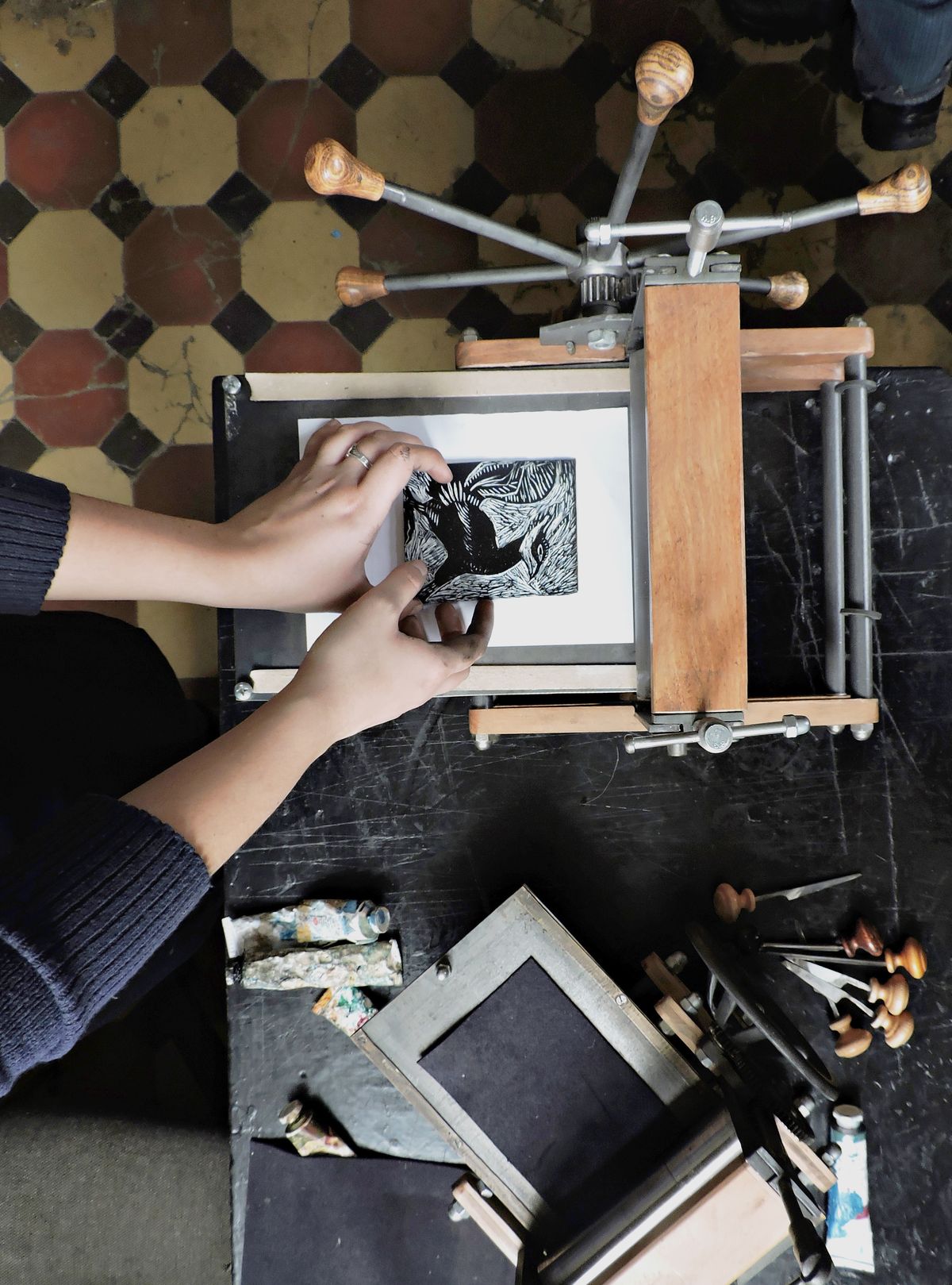 Printmaker's hands positioning a hand-carved linocut block on a manual tabletop press in the Legwork Press Grand Rapids studio